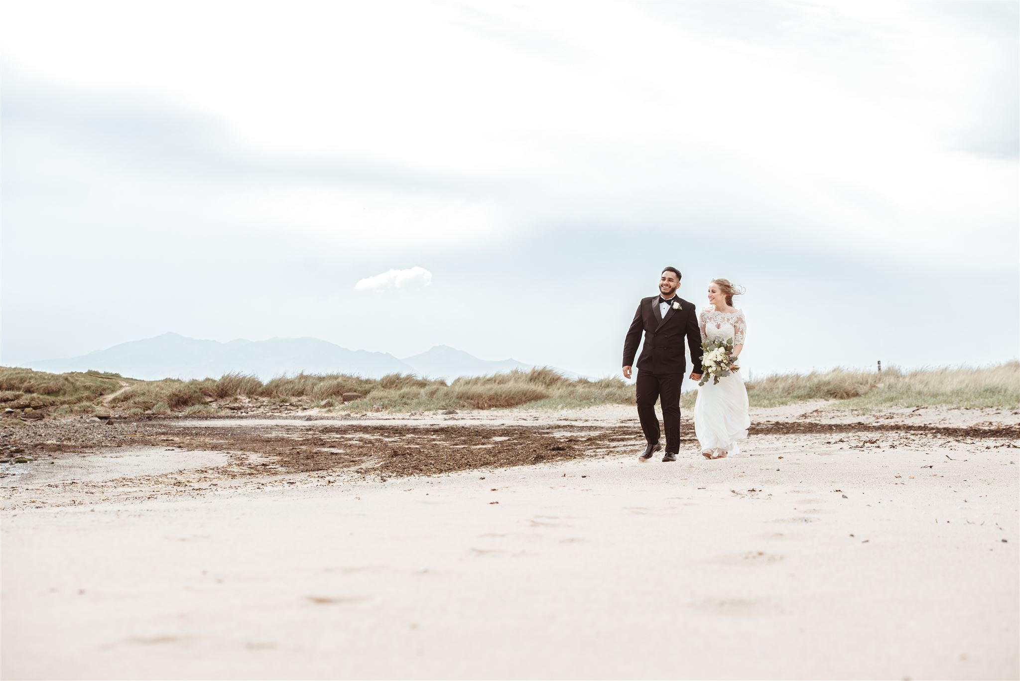 A bride and from walk hand in hand along a sandy beach. Grassy dunes can be seen in the distance and patches of seaweed lie to their right.